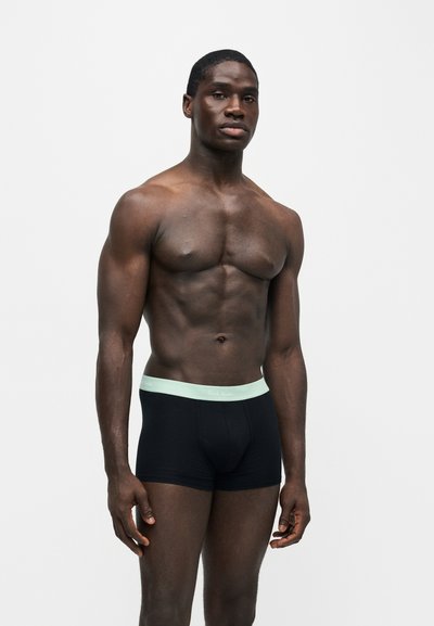 Homme musclé debout sur un fond blanc, portant un boxer noir avec une ceinture claire, regardant devant avec une expression neutre.