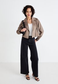 Jachetă din suede bej, scurtă, cu fermoar în față, blugi negri cu picioare largi și sandale negre cu bretele. Modelul poartă, de asemenea, un top alb cu dantelă la margine.