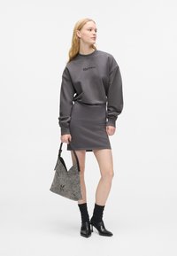 Kvinne iført grå langtermet kjole og svarte hæler, som holder en teksturert grå håndveske, stående mot en enkel hvit bakgrunn.