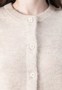 Lichtbeige cardigan gemaakt van zacht, gemêleerd stof. Heeft een ronde halslijn en vijf crèmekleurige knopen aan de voorkant. Eenvoudig en klassiek ontwerp.