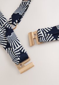 Gros plan de sangles en tissu à motifs bleu marine et blanc avec des fermoirs métalliques rectangulaires dorés, placées sur un fond beige clair.