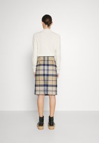 Beige wollen rok met een blauw ruitpatroon, tot op de knie, gecombineerd met een crème trui en zwarte enkellaarsjes op een lichtgrijze achtergrond.