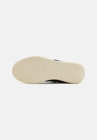 Black shoe sole featuring a textured light beige rubber material with "HUGO" embossed. Flat, rounded shape designed for comfort.