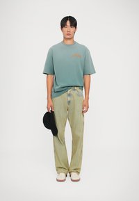 Lichtgroen oversized t-shirt met oranje tekst, gecombineerd met verticaal gestreepte lichtgele jeans. Witte sneakers en een zwarte pet in de hand.