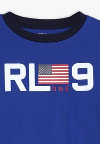 Modré tričko s námořnickým kulatým výstřihem. Na přední straně je bílé písmo "RL One" a "9", spolu s malou grafikou americké vlajky v červené a bílé.