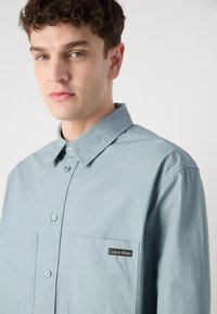 Šviesiai mėlyna marškinėlių palaidinė, pagaminta iš švelnaus ir lygus audinio, su apykakle, dviem priekiniais kišenėmis ir juodu Calvin Klein logotipo emblemą.
