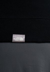Maglione nero con colletto a coste, caratterizzato da un'etichetta grigia cucita sul colletto che riporta la scritta "THE NORTH FACE" in bianco.