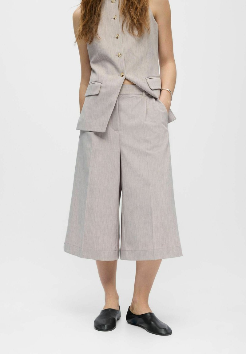 Femme portant un gilet sans manches gris clair boutonné avec un pantalon large court assorti et des chaussures noires sans lacets sur fond blanc.