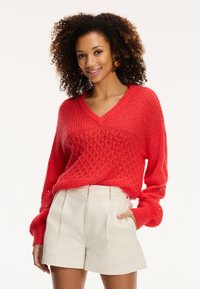Maglione rosso lavorato a maglia con scollo a V e intricato motivo a diamante, abbinato a pantaloni corti plissettati color crema.
