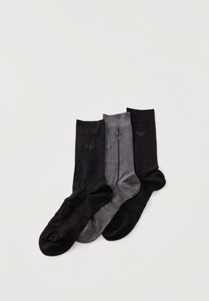 Trois chaussettes mi-mollet noires et grises, chacune ornée d’un petit logo d’aigle brodé, disposées sur un fond clair.