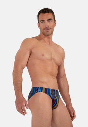 Muskulöser Mann stehend, trägt farbige gestreifte Slips, schaut zur Kamera mit neutralem Hintergrund.