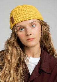 Żółta dziana czapka beanie o prążkowanej strukturze, z brązową skórzaną metką jako akcent; noszona przez modela z falującymi włosami i ciemną kurtką.