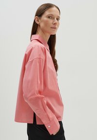 Chemise rose clair à manches longues, avec un col et des fentes sur les côtés. Le tissu semble lisse avec une texture subtile.