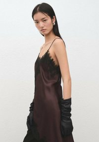 Vestido de satén burdeos con ribete de encaje negro, con tirantes finos y silueta ajustada al cuerpo. Combinado con guantes de cuero negros.