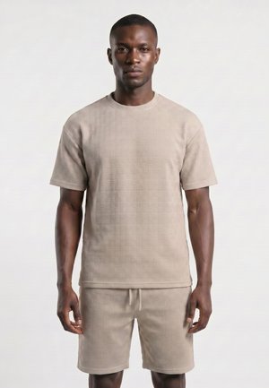 Homme portant une chemise beige texturée à manches courtes assortie et un short, debout devant un fond clair uni, regardant droit devant avec une expression neutre.