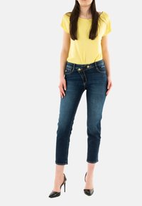 Jeans de mezclilla azul con un ajuste ajustado y largos hasta el tobillo, combinados con una camiseta amarilla holgada. Presenta cierre de doble botón y bolsillos frontales.