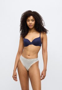 Soutien-gorge matelassé bleu marine avec un détail en nœud au centre. Culotte rayée beige et blanche avec bordure en dentelle. Tissus lisses et coupe ajustée.