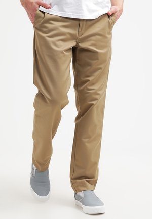 Man draagt beige chinos en grijze instapschoenen, met zijn handen in zijn zakken, lopend tegen een witte achtergrond.