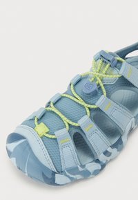 Ljust blå sandal med ovandel i mesh, gula detaljer, ett säkert snörsystem och en texturerad, mönstrad sula med rundad tå.
