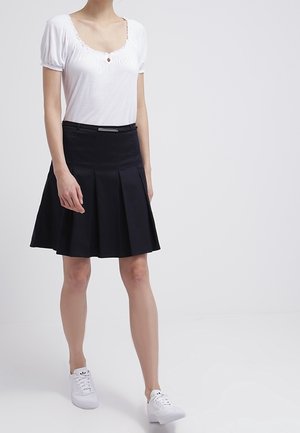 Femme portant un haut blanc à manches courtes, jupe plissée noire avec ceinture et baskets blanches, debout devant un fond clair uni.