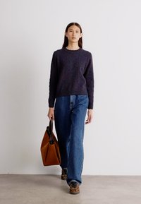 Marineblauer Strickpullover, lockere blaue Jeanshose und eine braune Umhängetasche. Gemusterte Schuhe sorgen für Textur. Das Modell geht vor einem schlichten Hintergrund.