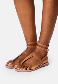 Kožené sandály v barvě tan s designem T-strap, tenkými popruhy kolem kotníku, plochou podrážkou a otevřenou špičkou, které vystihují hladkou texturu a minimalistický styl.