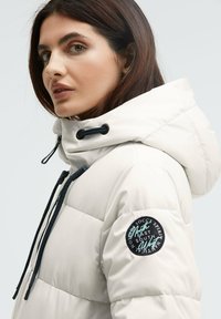 Weiße gepolsterte Jacke mit hohem Kragen und verstellbarem Kapuzenzug. Mit einem schwarzen runden Aufnäher am linken Ärmel, der Text- und Logodetails aufweist.