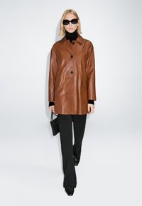 Blonde vrouw met zwarte zonnebril, bruine leren knopenjas, zwarte coltrui, zwarte broek, laarzen, en een kleine zwarte handtas.