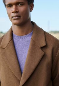 Brauner Wollmantel mit Reverskragen, getragen über einem helllila Strickpullover. Der strukturierte Stoff kontrastiert mit dem glatten Strick des Pullovers.