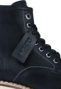 Bota de cuero negro hasta el tobillo con costuras blancas, ojales de metal y cordones negros. Presenta una etiqueta negra de marca adjunta al costado.