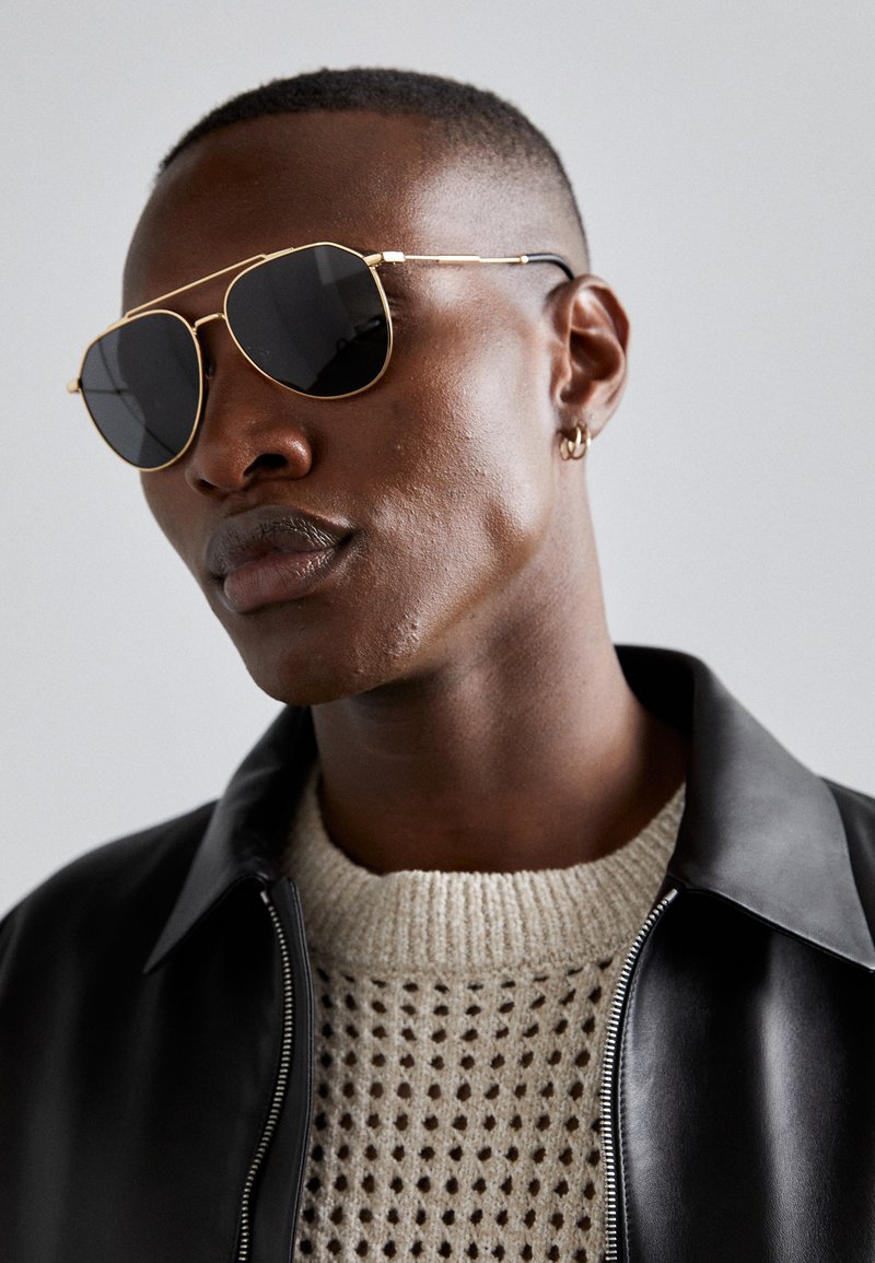 Laseikkarit, joissa on kultaiset kehykset ja tummat linssit, pukeutuneena neulottuun beigeen neuleeseen ja mustaan nahkatakkiin. Sileä pinta ja terävä muoto.