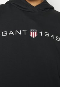 Černá mikina s kapucí s texturovaným povrchem, s bílým nápisem "GANT 1949" a štítovým logem s červenými a modrými pruhy.