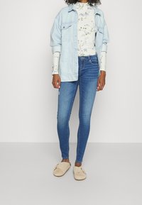 Chaqueta de denim azul claro sobre un cuello de tortuga blanco con estampado floral y jeans ajustados azules, combinada con zapatos tipo slip-on beige.