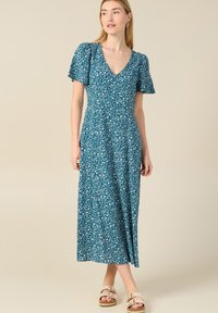 Tee-farbene Maxikleid mit floralen Mustern, kurzen Flatterärmeln, V-Ausschnitt und einem fließenden Schnitt. Stylisch kombiniert mit beigen Sandalen für einen lässigen Look.