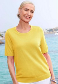 Gelber kurzärmliger Pullover aus weichem Strickmaterial, mit einem Rundhalsausschnitt und einer lockeren Passform. Getragen mit weißen Hosen.