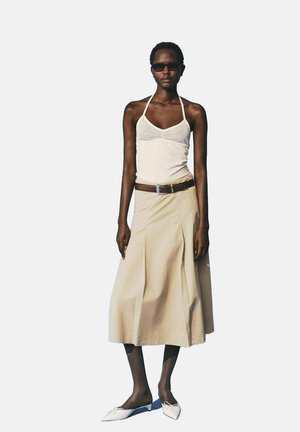 Vrouw in een witte haltertop, beige midi-jurk met een bruine riem, witte puntige flats en donkere zonnebril, staand voor een effen achtergrond.