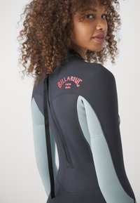 Dräkt med svart kropp och ljusblå ärmar. Har en logotyp på baksidan och en svart dragkedja. Materialet verkar vara elastiskt och texturerat.