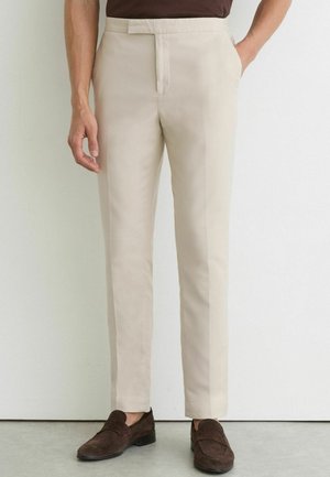 Homme portant un pantalon beige élégant avec poches latérales et mocassins en daim marron, debout contre un mur blanc.