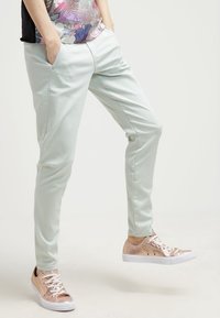 Světlé mátové kalhoty s úzkým střihem, s bočními kapsami a hladkou strukturou, v kombinaci s růžově zlatými teniskami a květinovým topem.