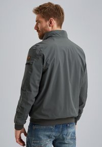 Chaqueta bomber gris con cuello alto, cierre frontal con cremallera y puños elásticos; presenta un parche en la manga izquierda y una textura suave y mate.