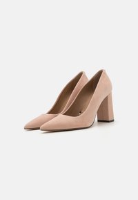 Beige velúr magassarkú pumps, hegyes orral és vastag sarokkal. Simaság jellemzi, letisztult dizájn, minimális részletek és fekete talp.