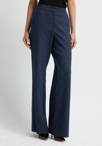 Vrouw in marineblauwe pinstripe wijde broek en zwarte puntige hoge hakken, staand tegen een effen achtergrond.