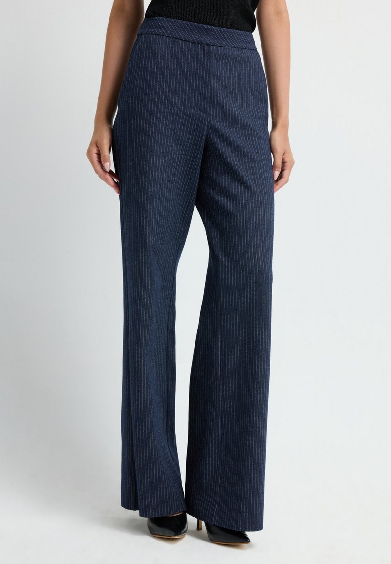 Vrouw in marineblauwe pinstripe wijde broek en zwarte puntige hoge hakken, staand tegen een effen achtergrond.
