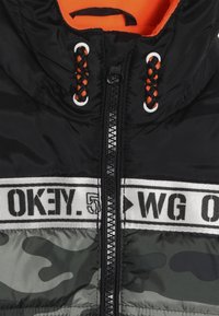 Chaqueta acolchada con capucha negra, forro naranja, cremallera frontal y un patrón de camuflaje gris en la parte inferior con una banda de texto en contraste.