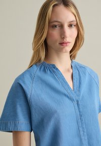 Camicia in denim azzurro chiaro, con maniche corte, scollo a V e dettagli di cucitura sottili. Texture liscia, design casual.