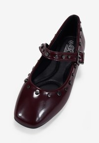 Zapato plano tipo Mary Jane de charol burdeos con punta cuadrada, detalles de tachuelas y una correa ajustable con una hebilla metálica.