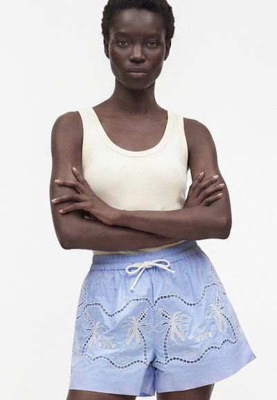 Femme, bras croisés, portant un haut blanc sans manches et un short bleu clair avec des motifs de palmiers brodés blancs.