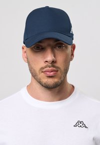 Navy blå caps laget av stoff, med en buet skjerm og en logo aksent. Båret med en hvit t-skjorte som viser en liten svart logo.