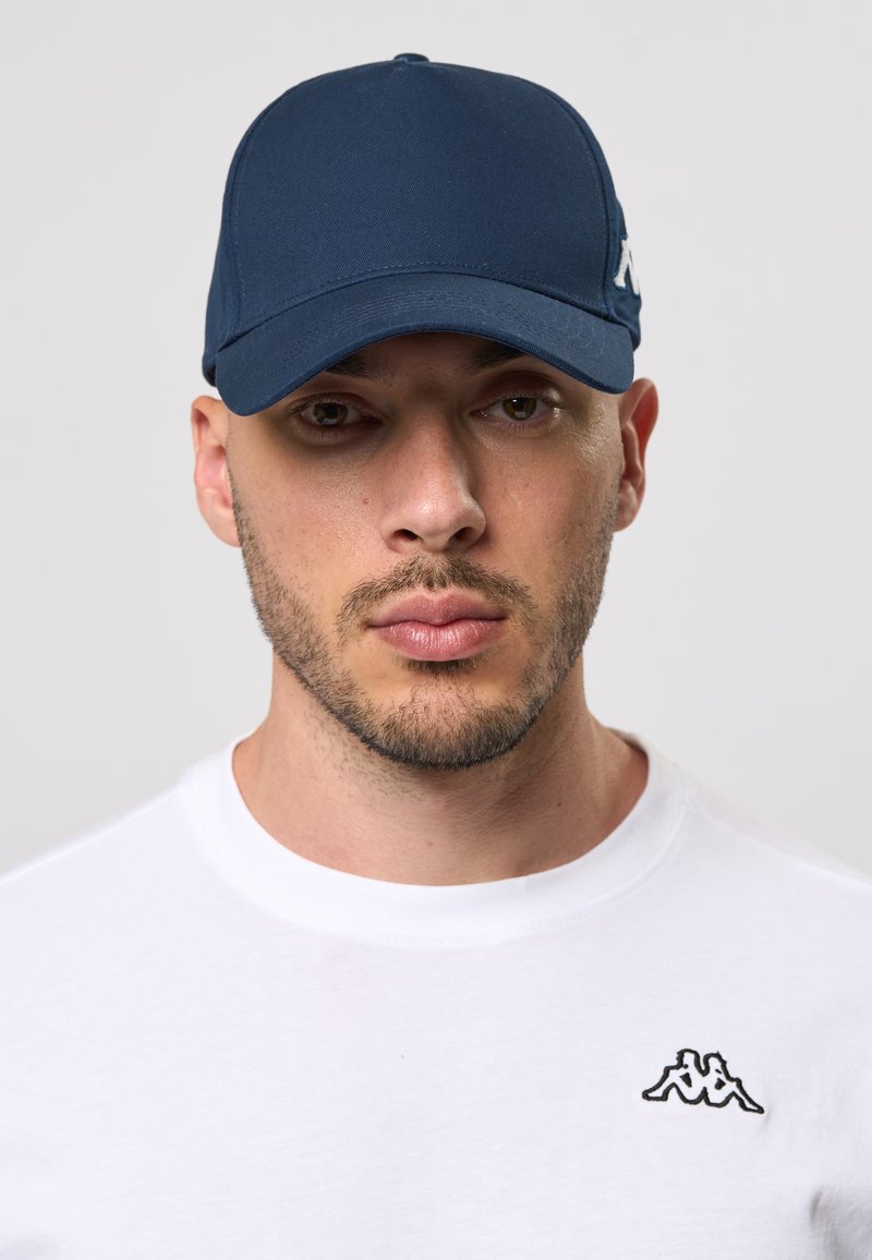 Navy blå caps laget av stoff, med en buet skjerm og en logo aksent. Båret med en hvit t-skjorte som viser en liten svart logo.