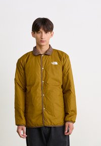 Bruine geïsoleerde jas met een contrasterende bruine kraag. Voorzien van drukknoopsluitingen en een klein wit logo op de linkerborst. Zachte stoftextuur.